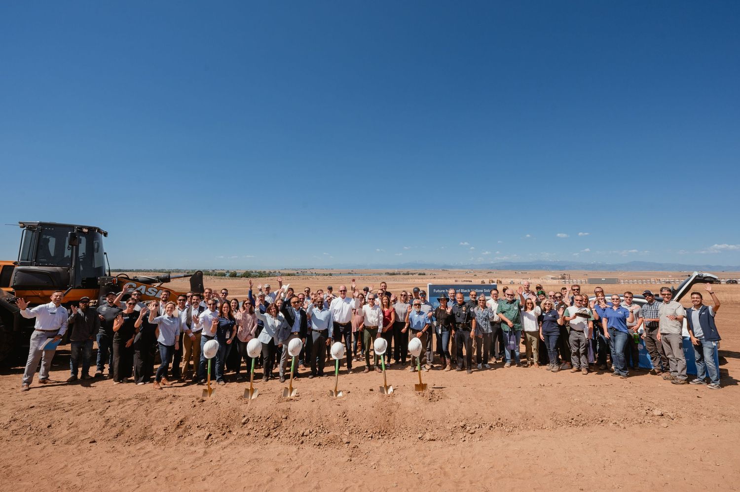 Large group of people gathered outdoors on dusty ground with farm equipment and blue sky in background.