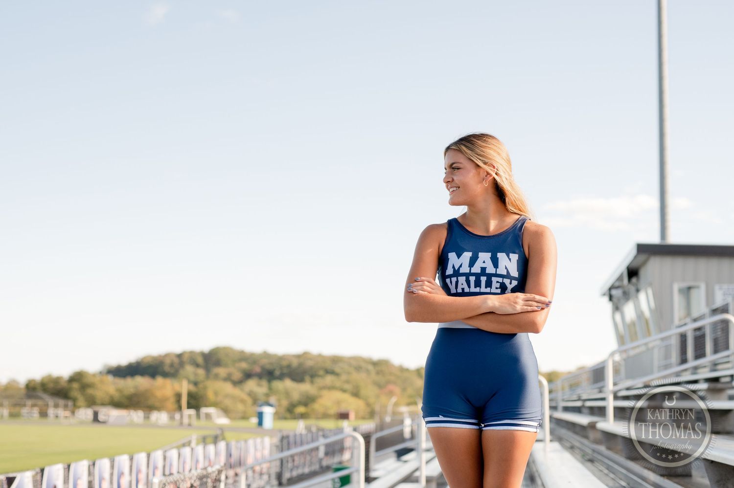 Athletic person in blue uniform stands confidently on bleachers against scenic backdrop.