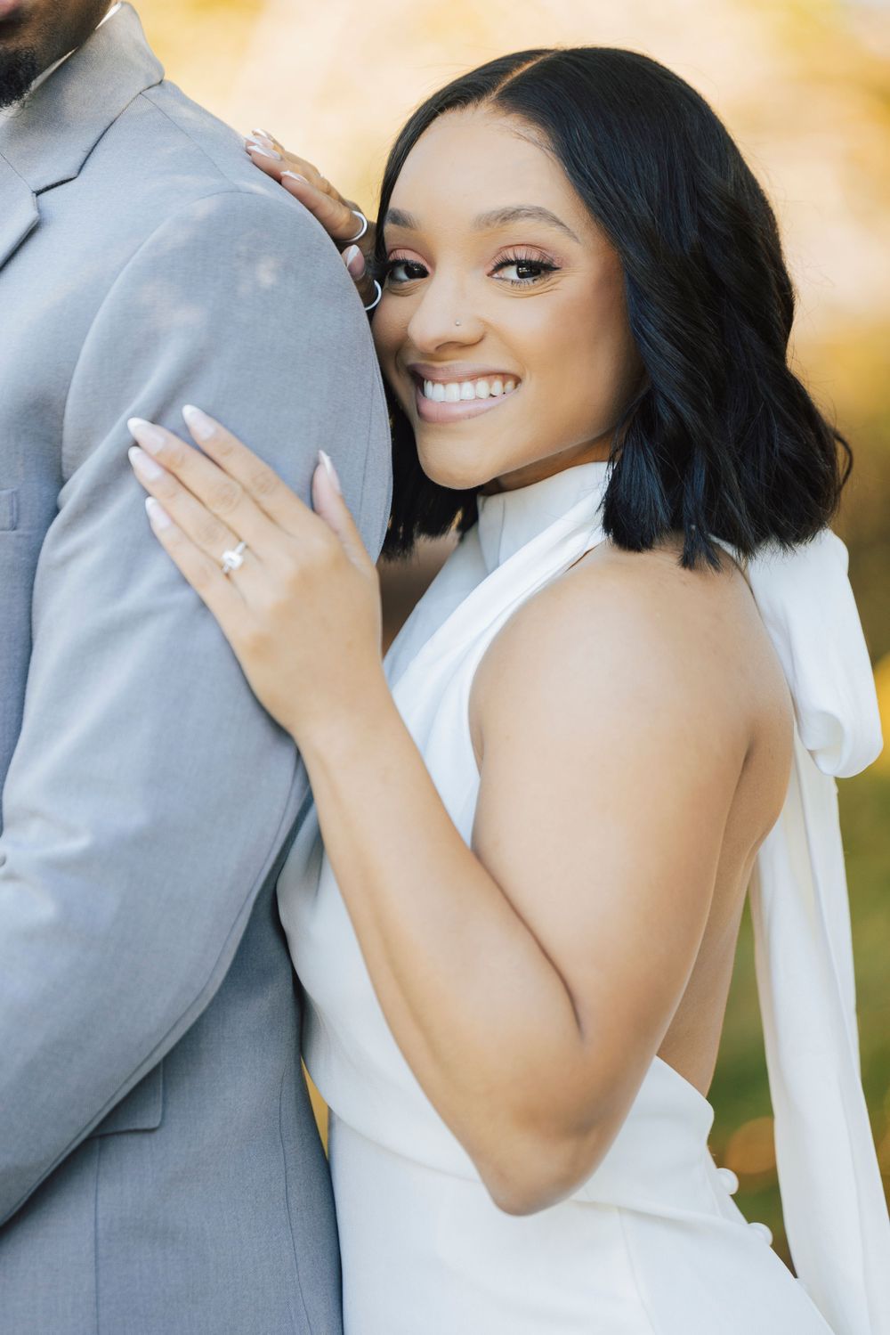 A bride in a white dress shows off her engagement ring while embracing her partner in a grey suit outdoors.