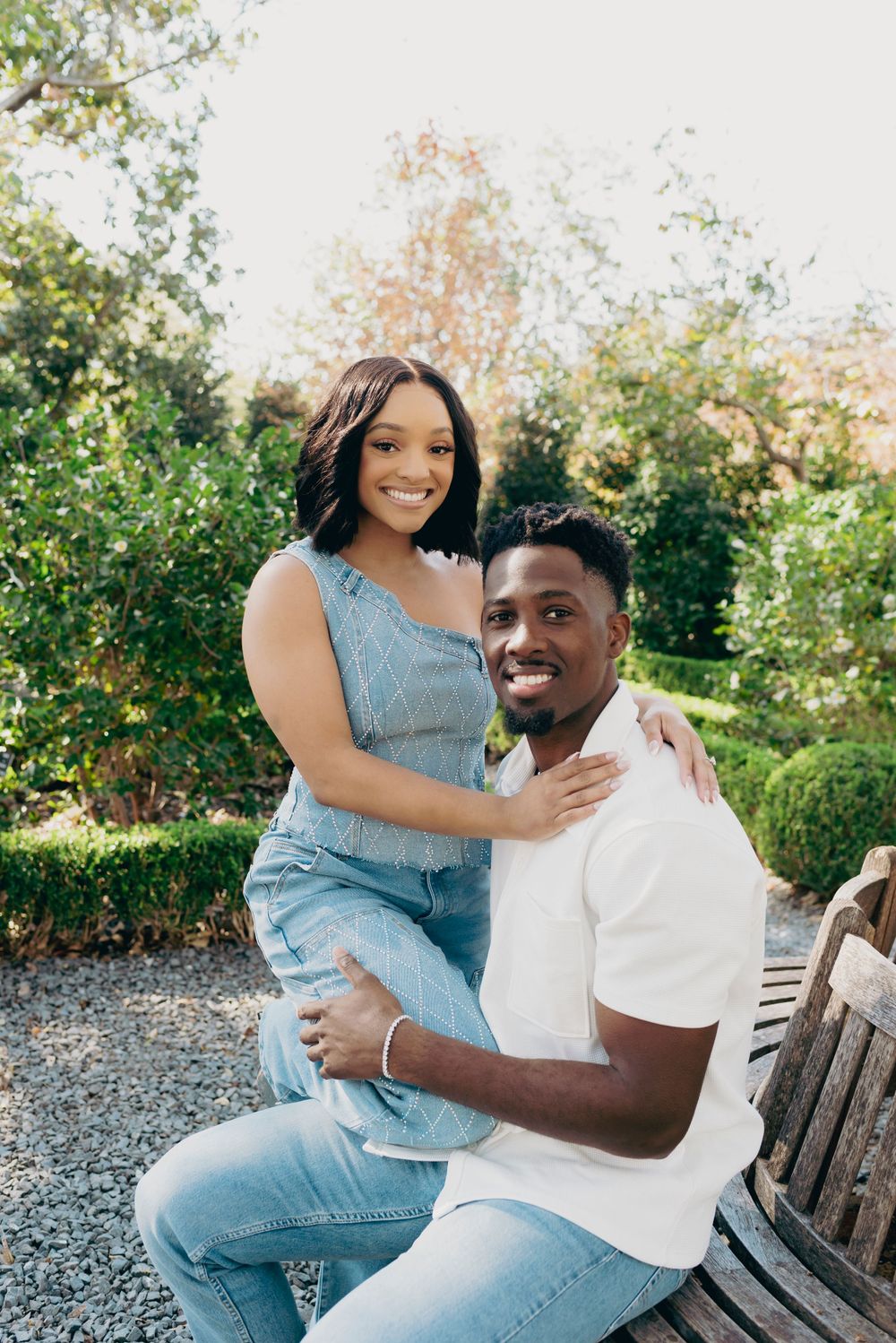 A happy couple embraces while sitting on a wooden bench in a lush garden setting on a sunny day.