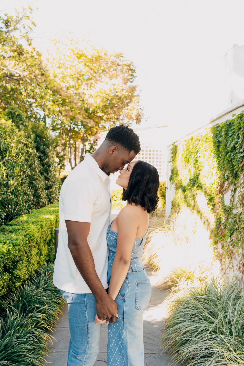 A romantic couple holds hands and shares an intimate moment in a sunlit garden pathway surrounded by greenery.