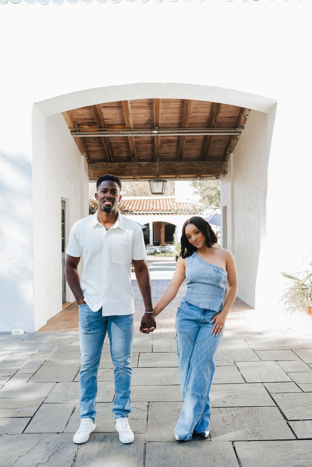 A couple wearing coordinated blue and white outfits hold hands while standing under an arched walkway.