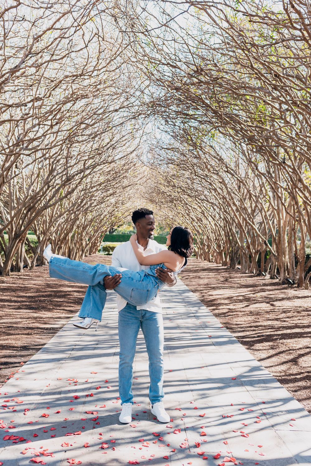 Romantic couple sharing a sweet moment under an archway of bare tree branches with flower petals scattered on the path.