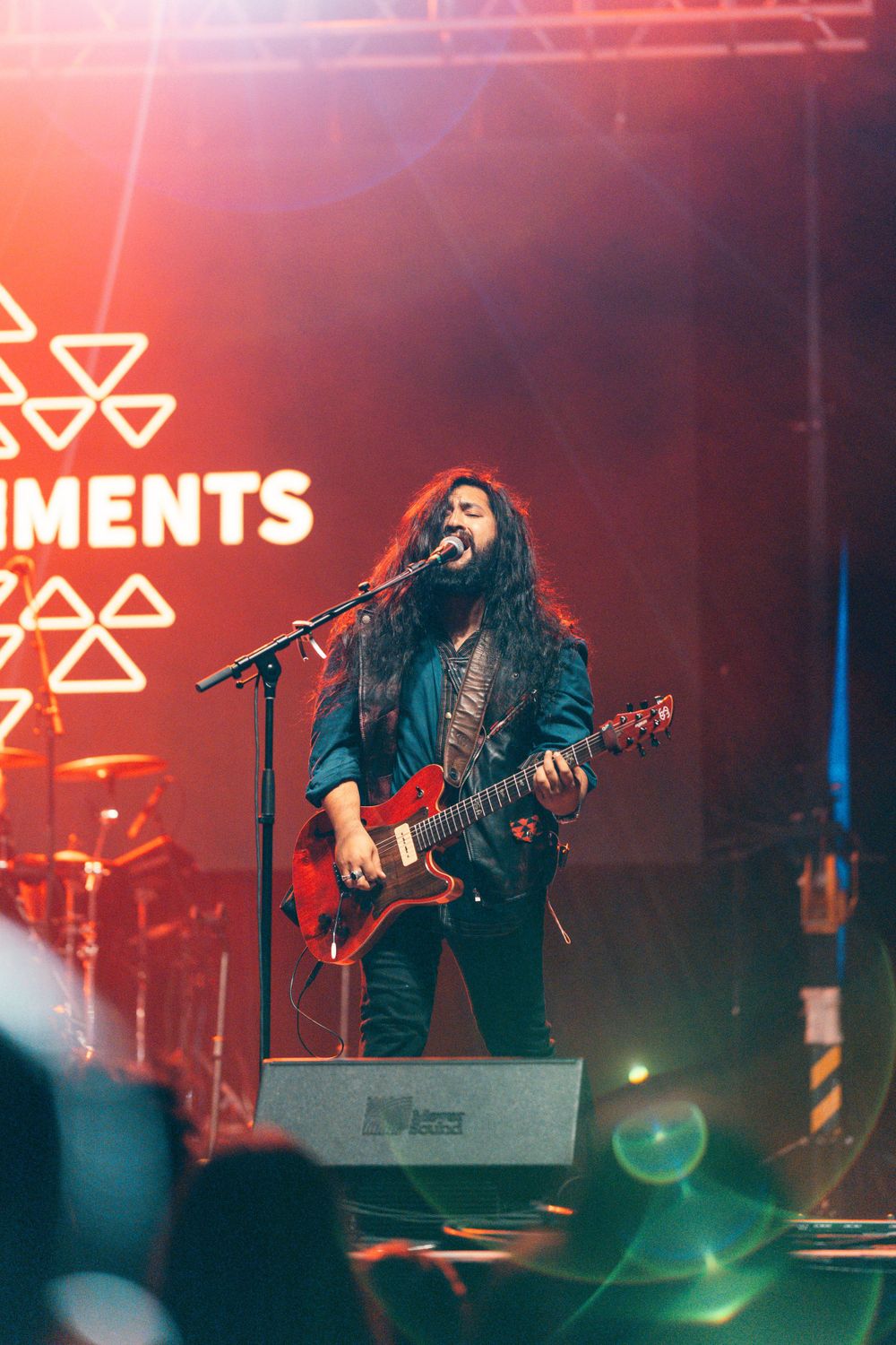 A guitarist performs on stage under red lighting with The Elements band logo visible.