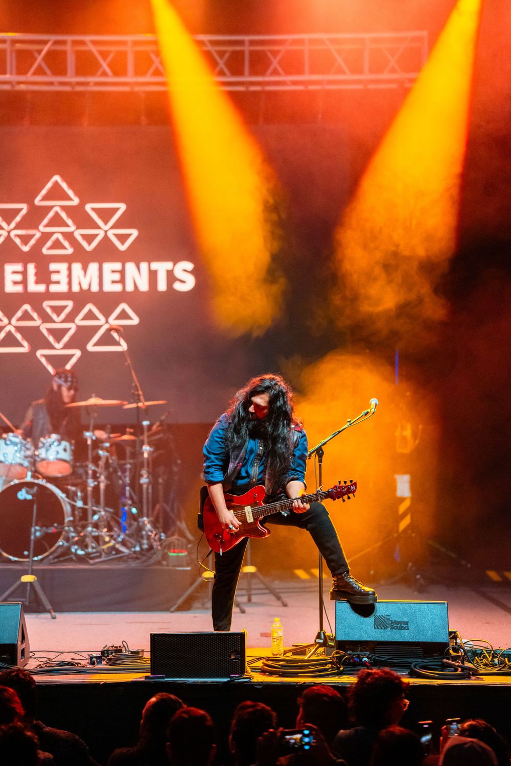 A performer plays guitar energetically on stage with orange lighting and The Elements logo.