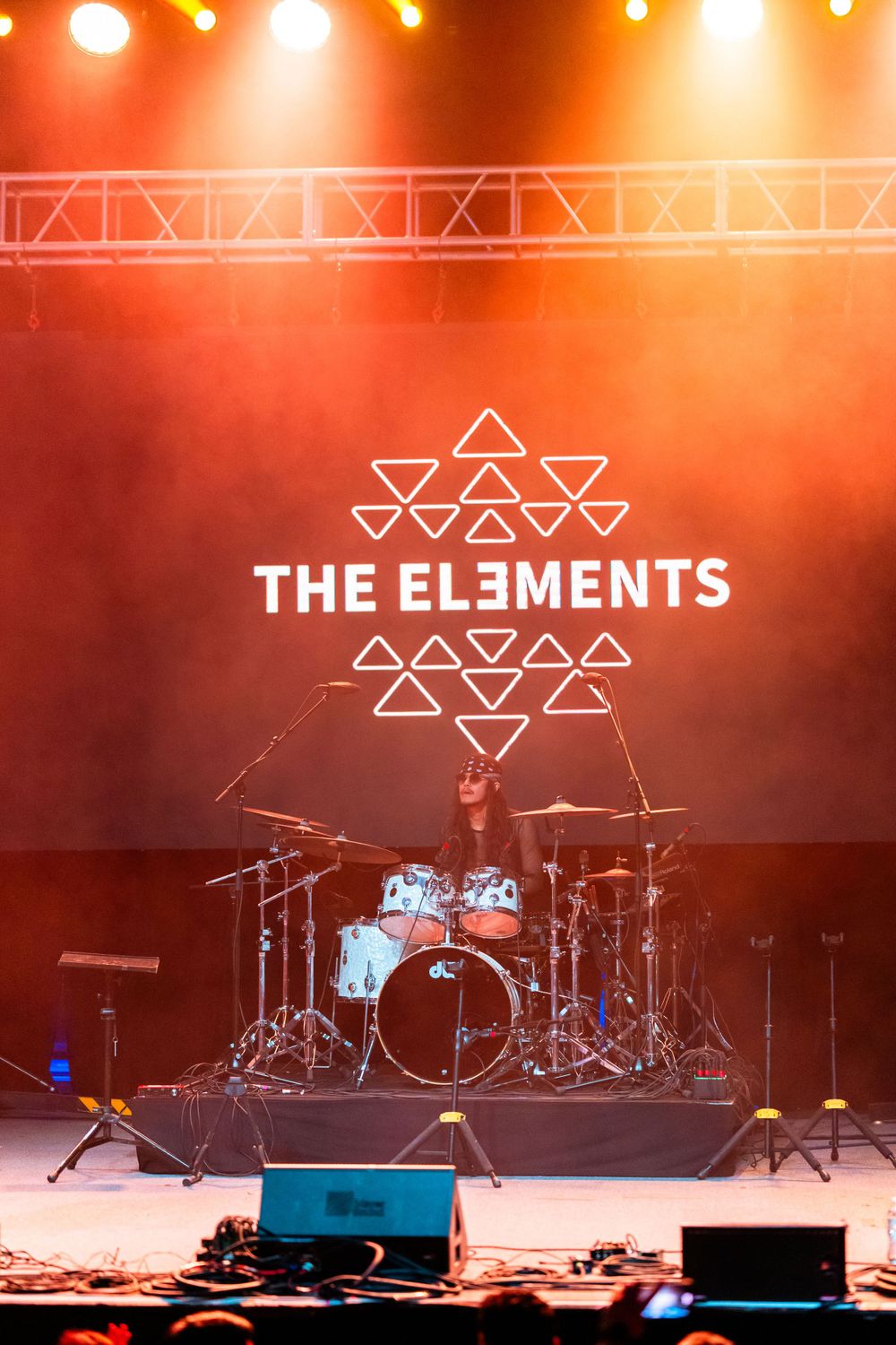 A drum kit on stage with 'The Elements' logo display featuring geometric triangles and stage lighting in orange tones.