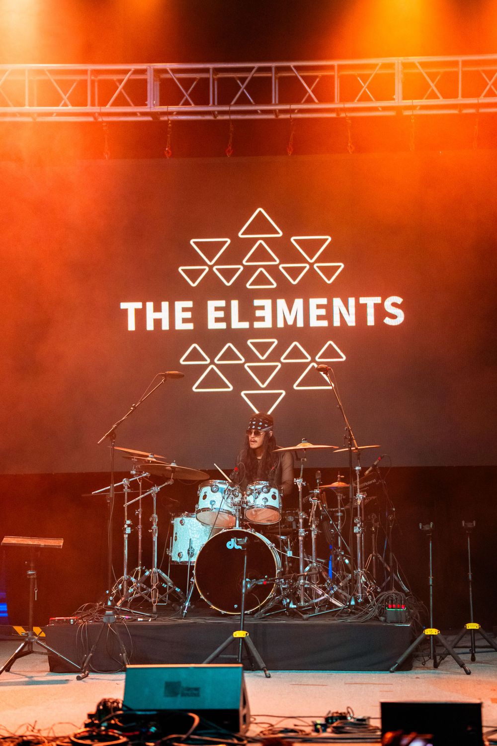 Empty concert stage setup with drum kit and 'The Elements' triangular logo illuminated in warm lighting.
