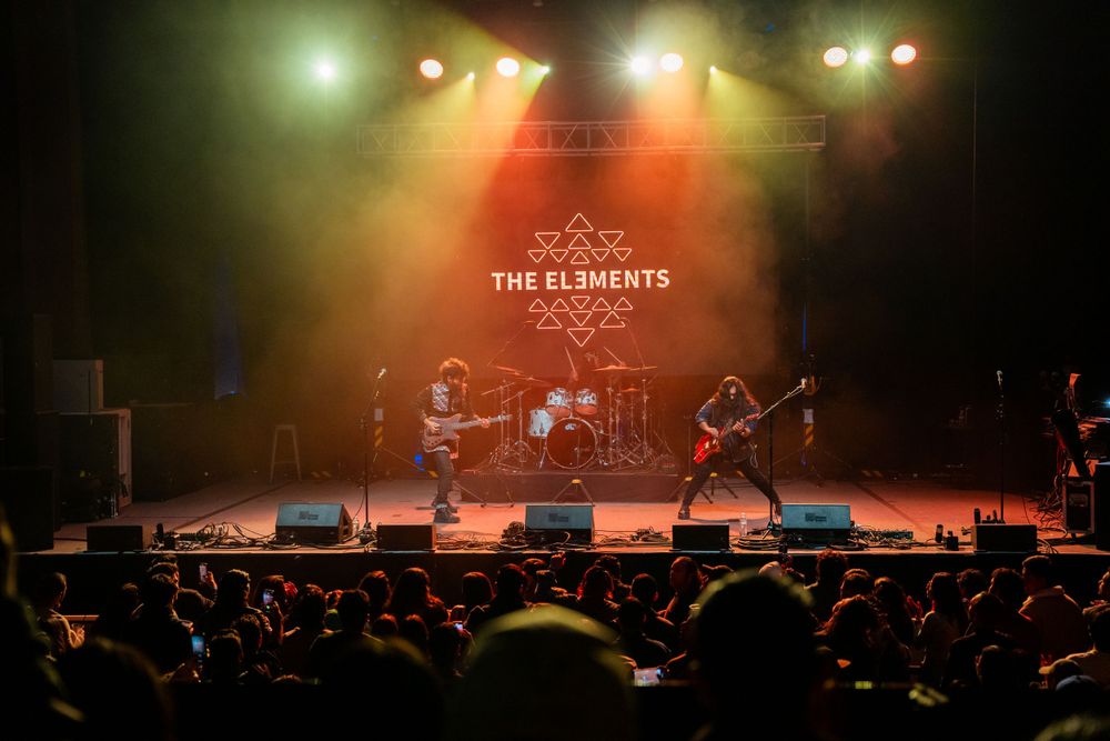Wide shot of rock band performing on stage with crowd silhouettes visible in foreground.