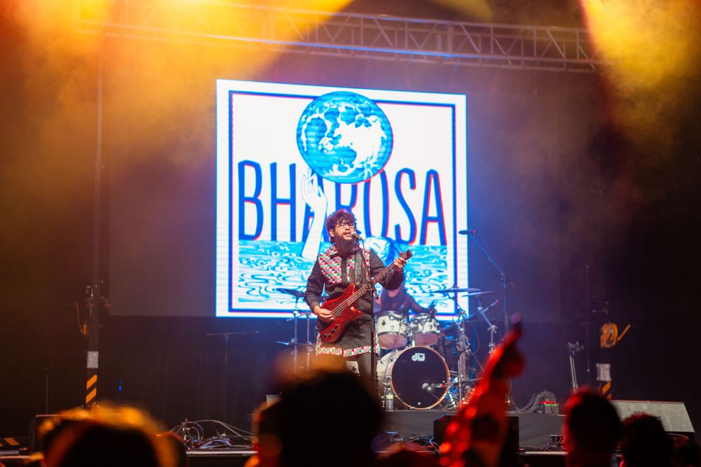 Concert performance with 'Bhatoosa' logo projected on screen behind band in warm lighting.