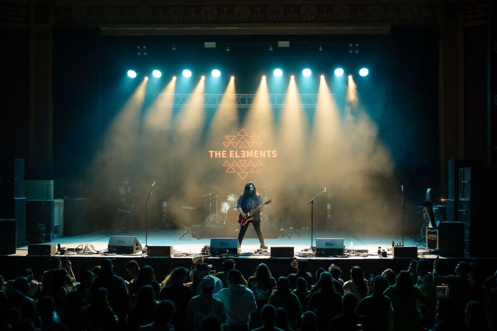 Concert venue with bright white stage lights illuminating performers through atmospheric haze.