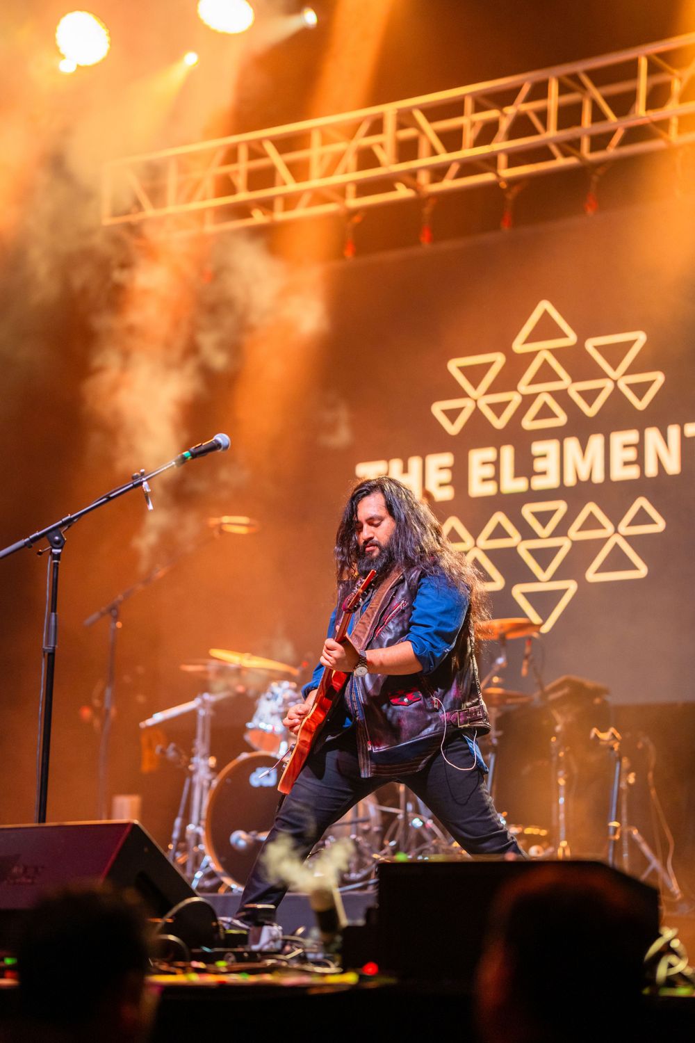 Guitarist performing on stage under 'The Elements' geometric logo with orange lighting and fog effects.