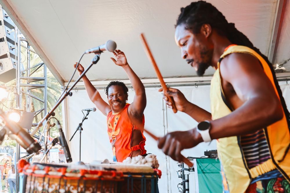 Two musicians perform African drumming on stage at an outdoor festival, wearing colorful traditional attire.