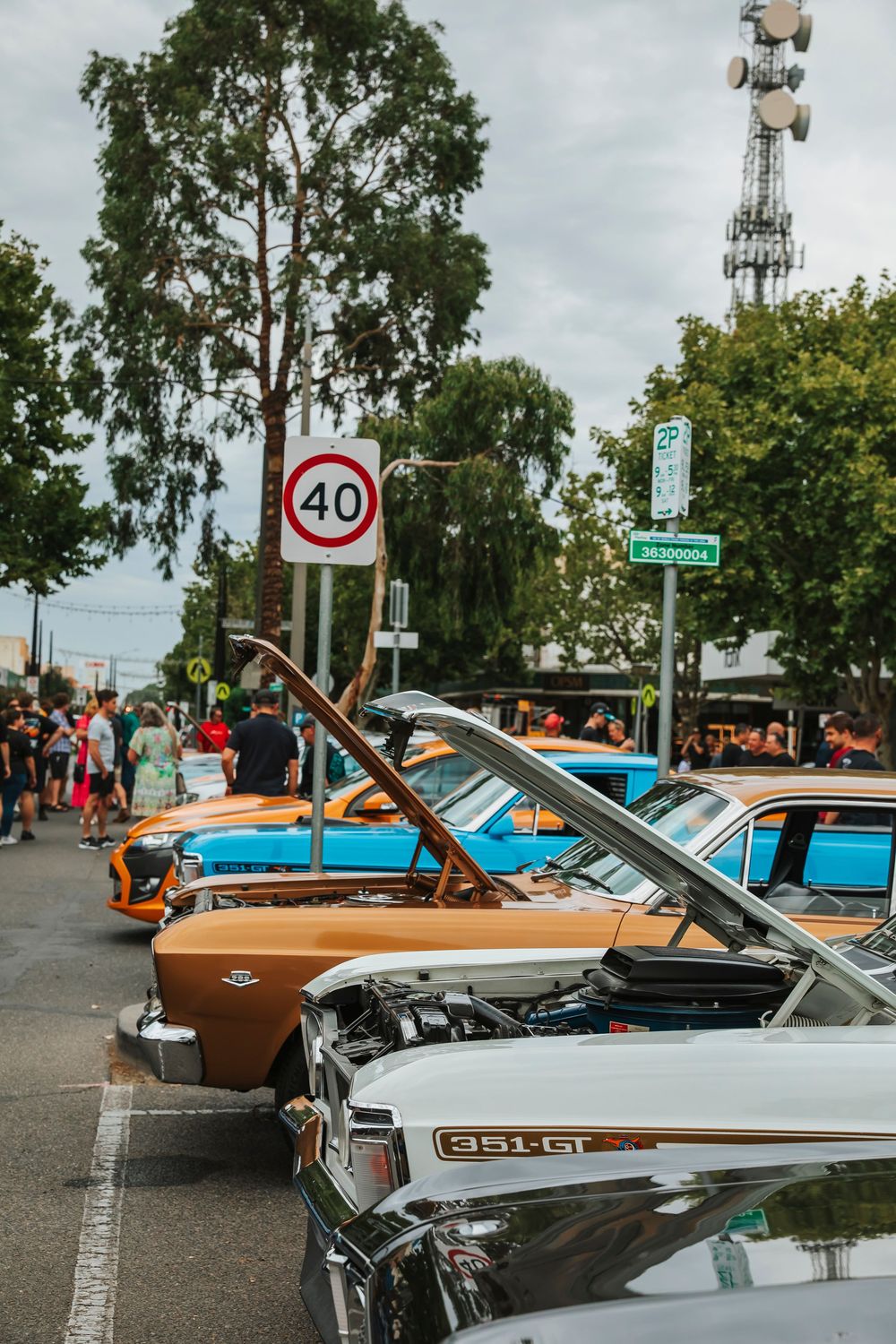 Classic cars with open hoods on display at a car show against a cloudy sky background with a speed limit sign visible.