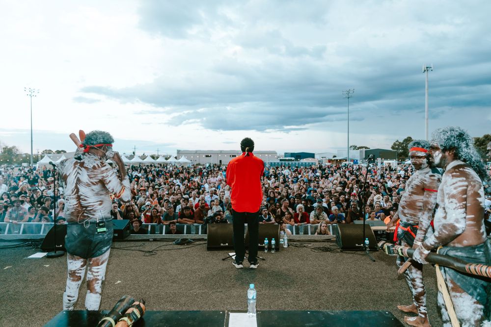 A performer stands on stage flanked by band members in front of a large festival crowd.
