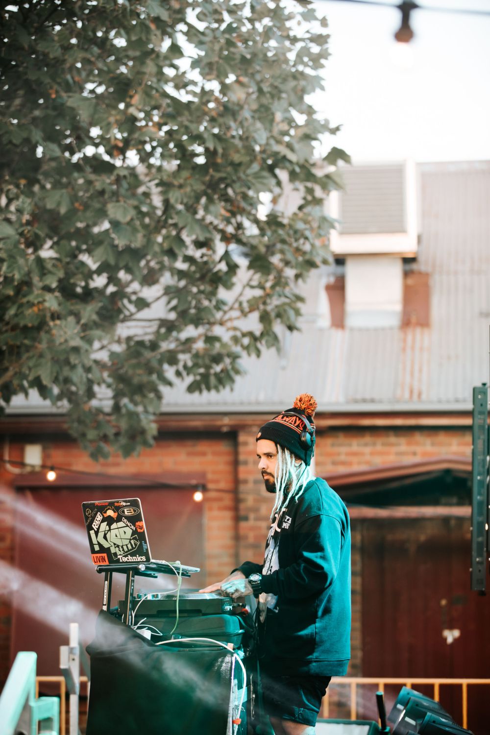 DJ performing at outdoor venue with turntables wearing green hoodie and winter hat.