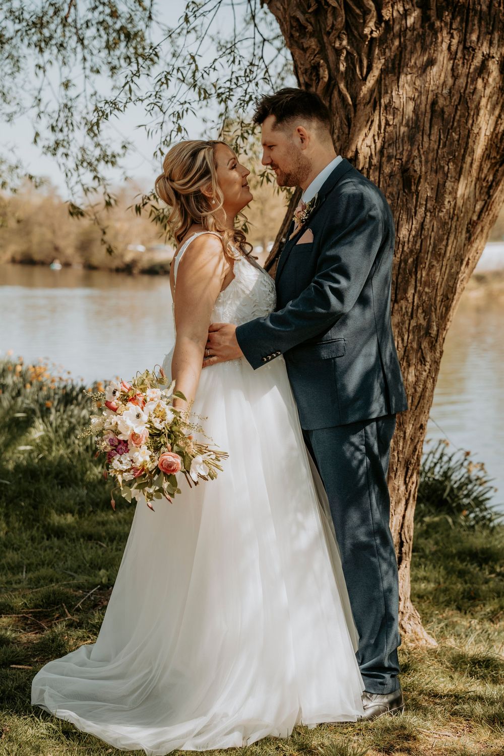 A romantic wedding couple embraces under a tree by a lake, with the bride holding a lovely pink and white bouquet.