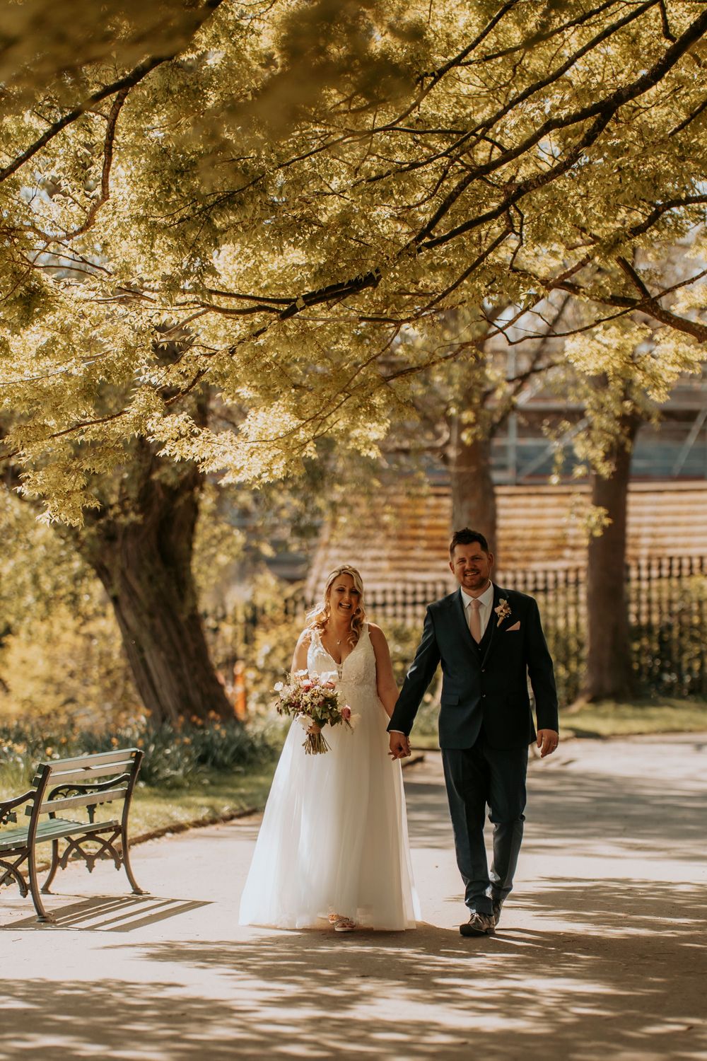 A newlywed couple walks together on a sunlit path under golden autumn trees at their outdoor wedding ceremony.