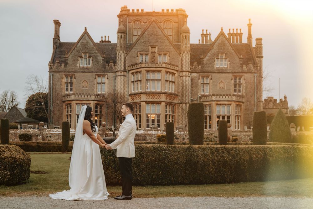 A romantic wedding couple poses at sunset in front of a grand Victorian Gothic mansion with beautiful landscaping.