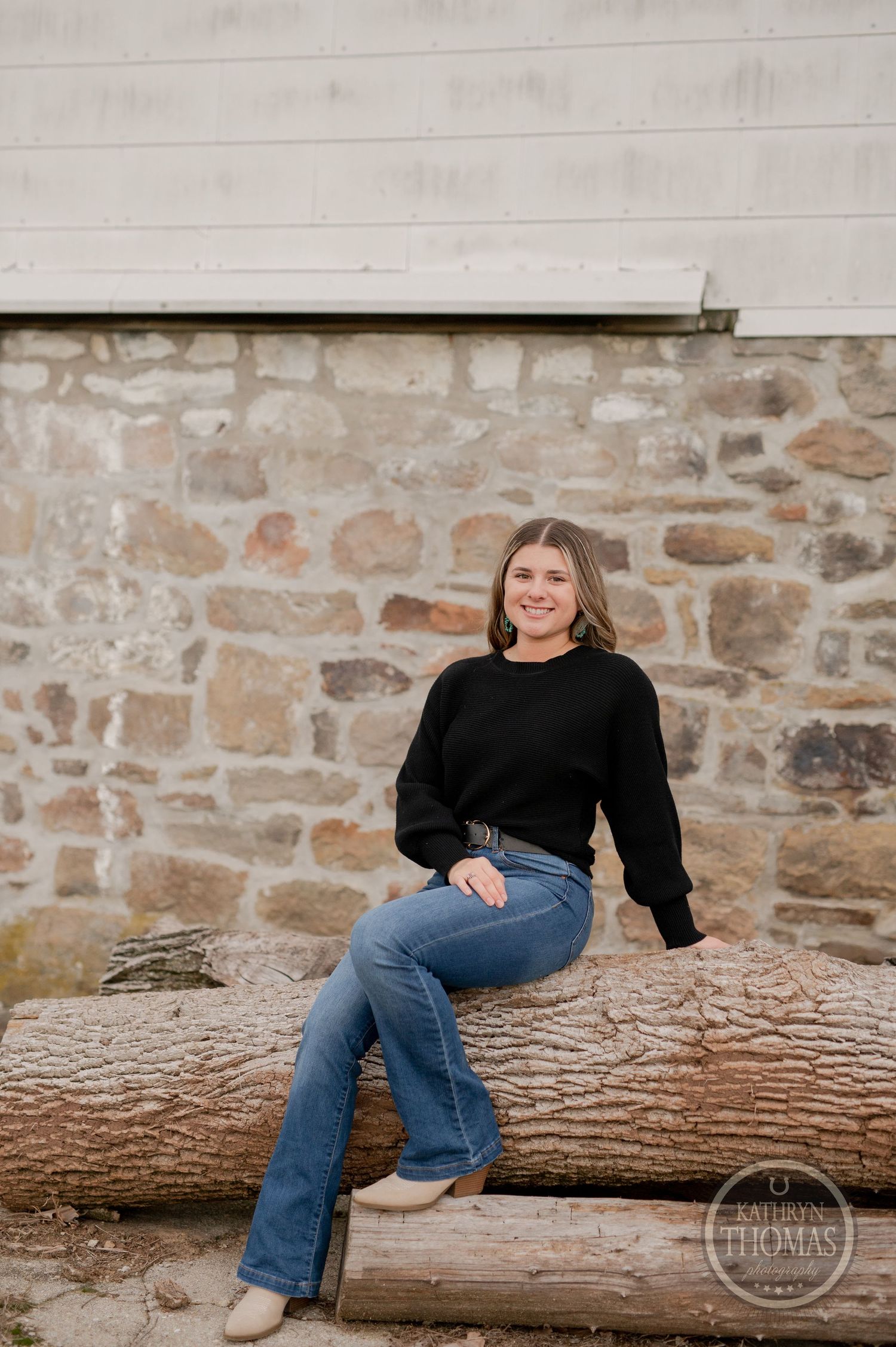 Natural outdoor portrait sitting on log against stone wall wearing casual black sweater and jeans.