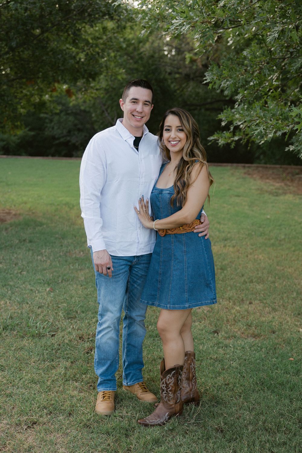 A couple in casual attire poses together on green grass during an outdoor engagement photo session.