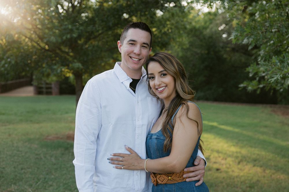 A couple in coordinated white and blue outfits embraces outdoors during a golden hour photoshoot in a grassy field.