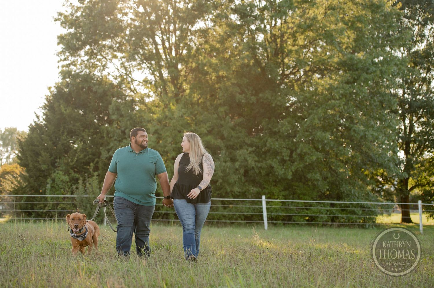 A couple walks with their brown dog through a grassy field at sunset.