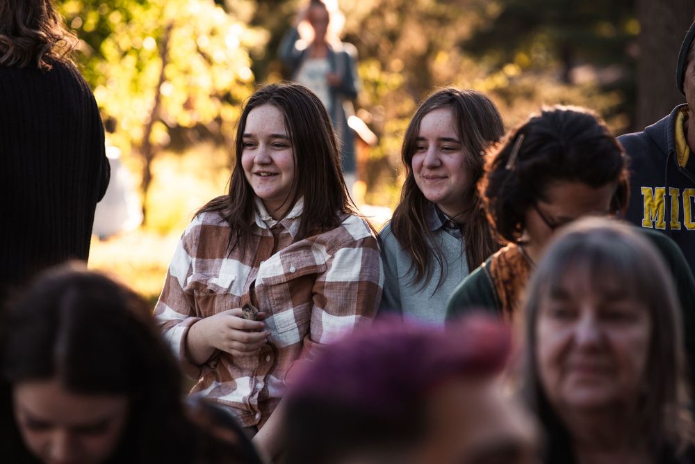 A candid crowd scene at an outdoor gathering with filtered sunlight streaming through trees in the background.