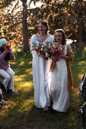Two brides in elegant white dresses walk together holding vibrant bouquets at an outdoor sunset wedding celebration.