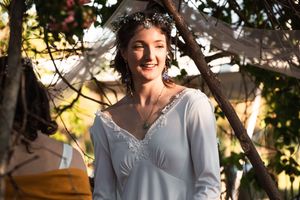 A person wearing a white lace dress and floral crown smiles radiantly in golden sunlit garden setting.