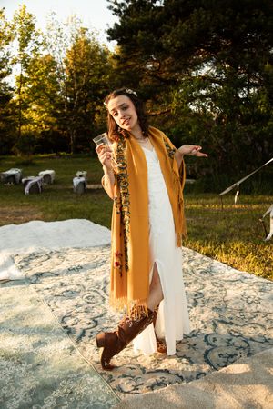 A person in a white dress and mustard yellow shawl poses outdoors on a patterned rug at sunset with sheep in background.