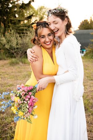 Two people embrace and laugh joyfully outdoors, one wearing a bright yellow dress and one in a white dress with floral bouquet.