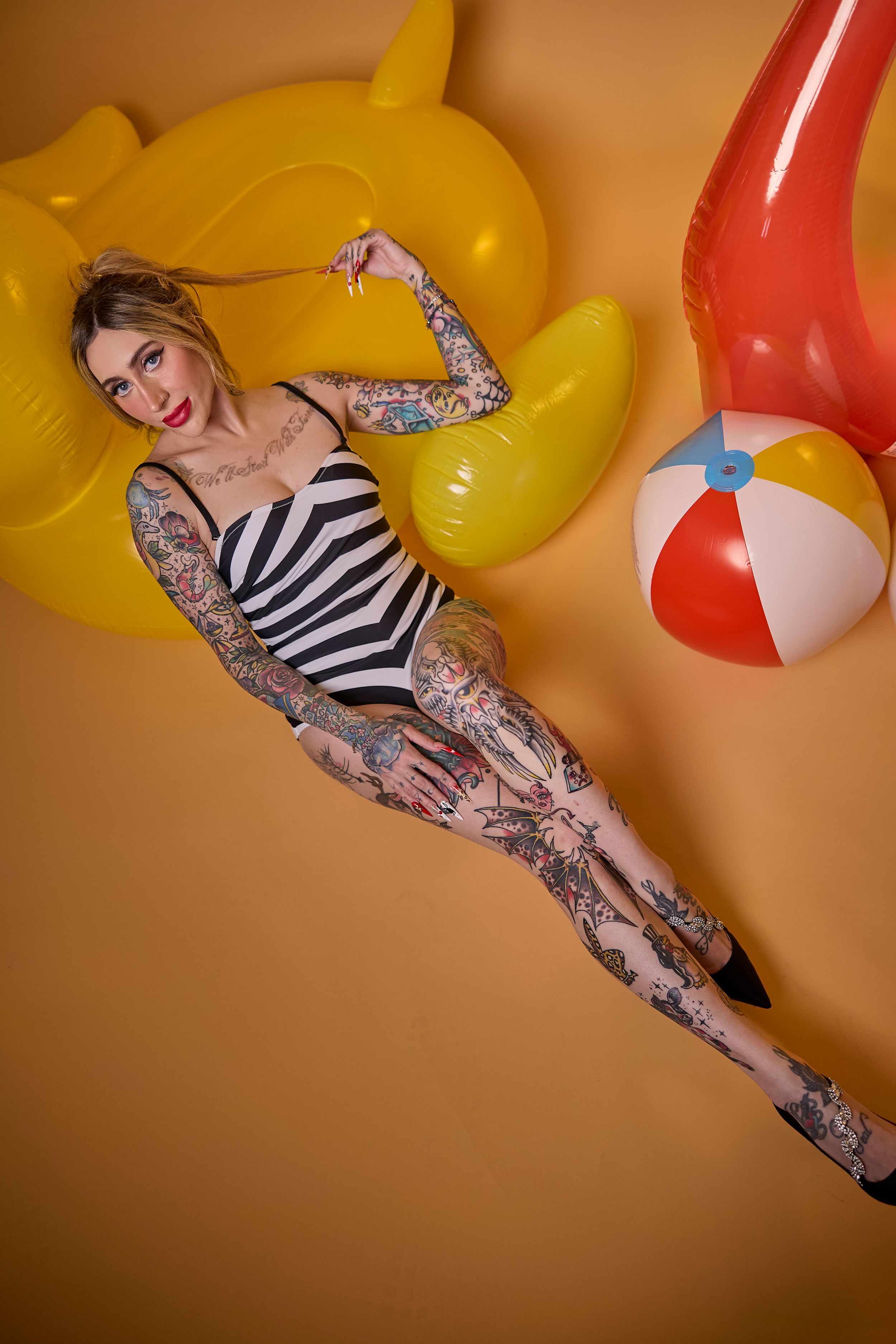 Model in striped swimsuit posing with yellow inflatable duck and beach ball against orange backdrop.