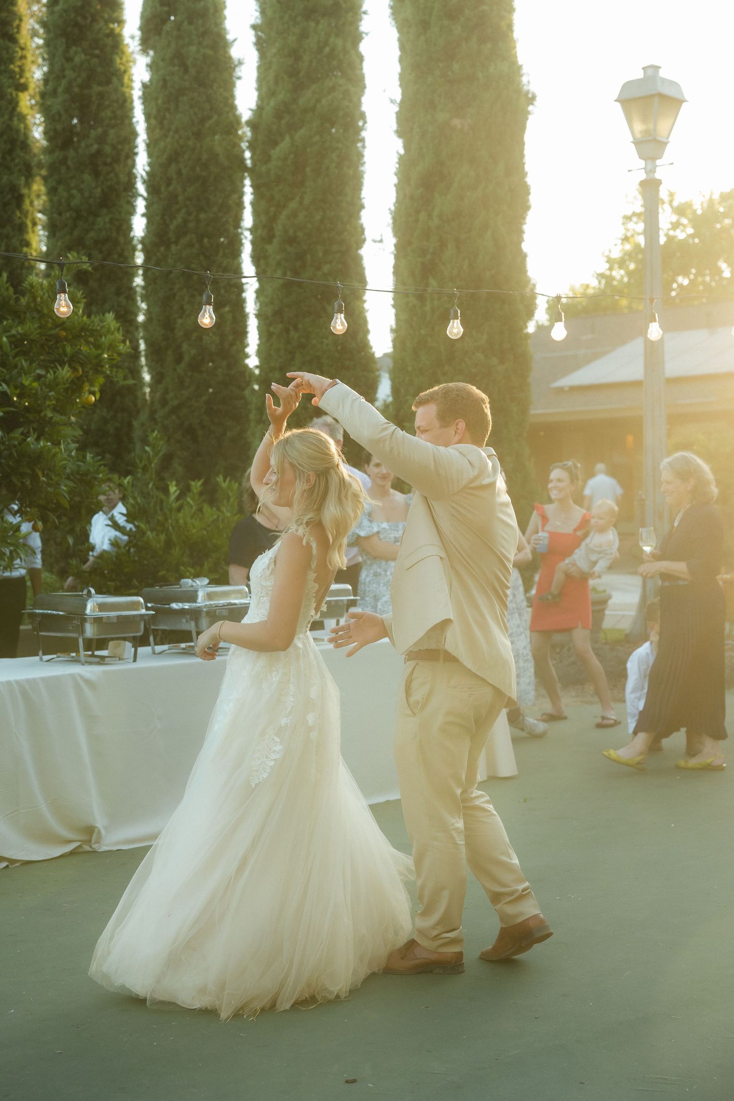A couple dances at their wedding reception with tall cypress trees and string lights in the background.