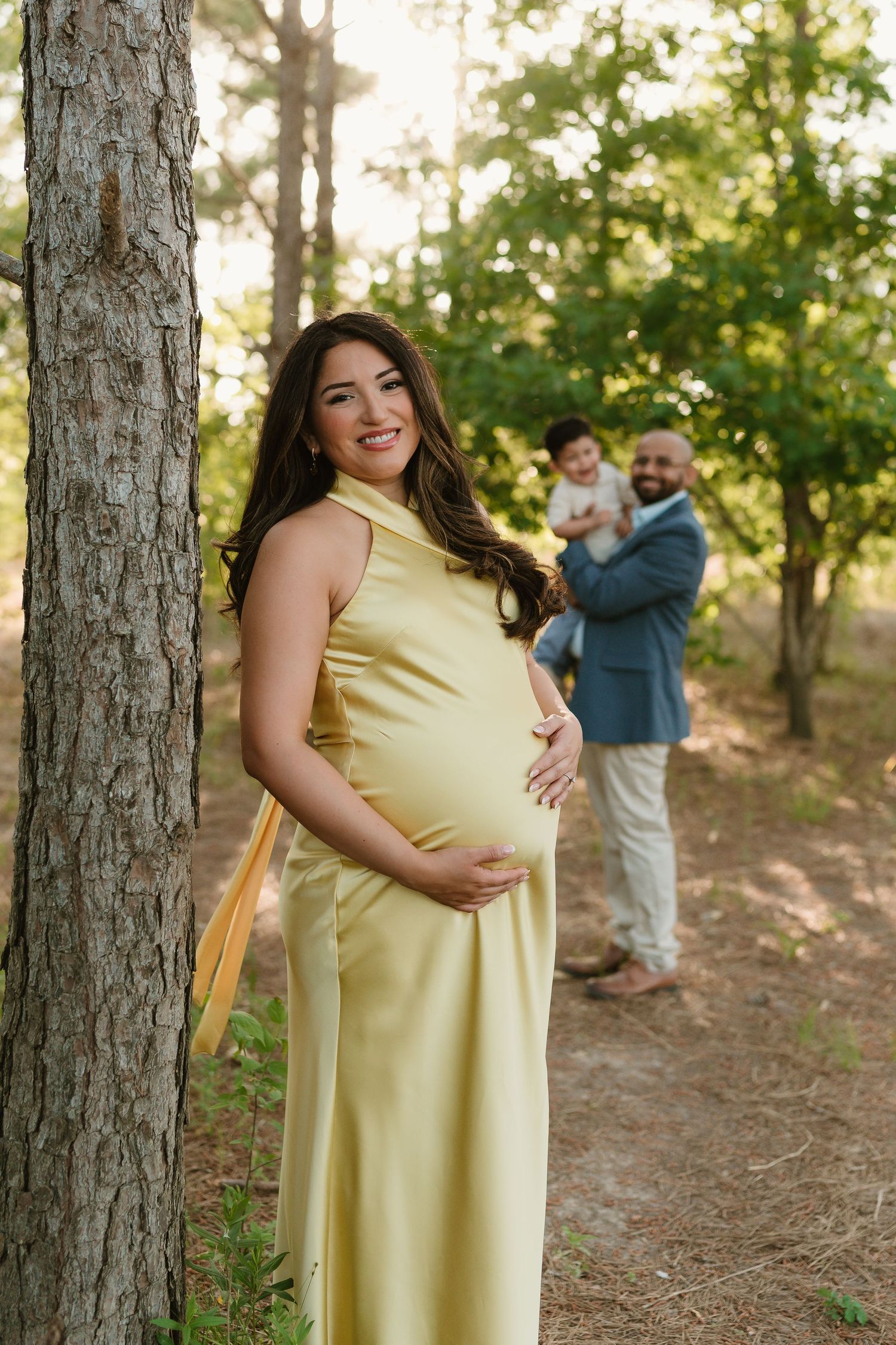 Maternity photos in a wooded setting with a woman in a yellow dress among pine trees.