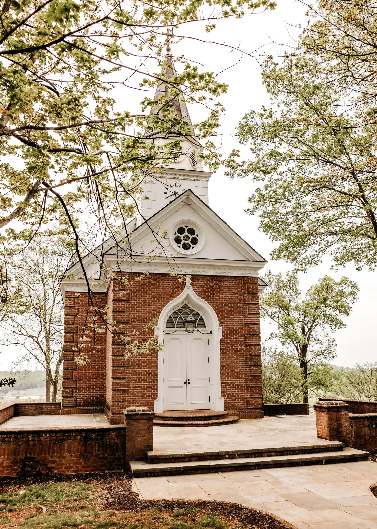 A small brick chapel with white Gothic arched doorway and steeple framed by spring dogwood blossoms on a misty day.