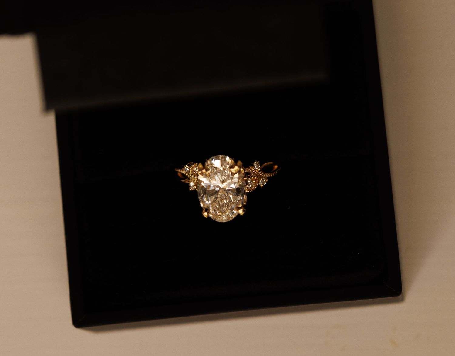 A vintage-style gold engagement ring with a large center diamond and ornate floral design details rests in a black display box.
