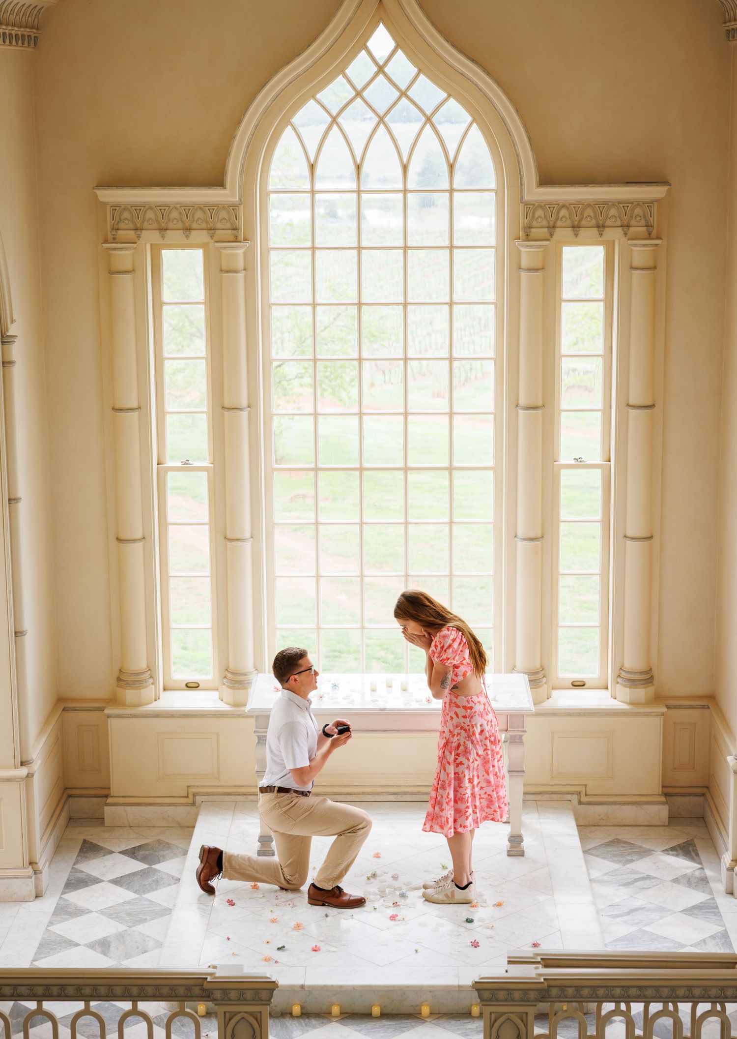 A romantic proposal moment in front of a grand Gothic arched window with rose petals scattered on a marble floor.