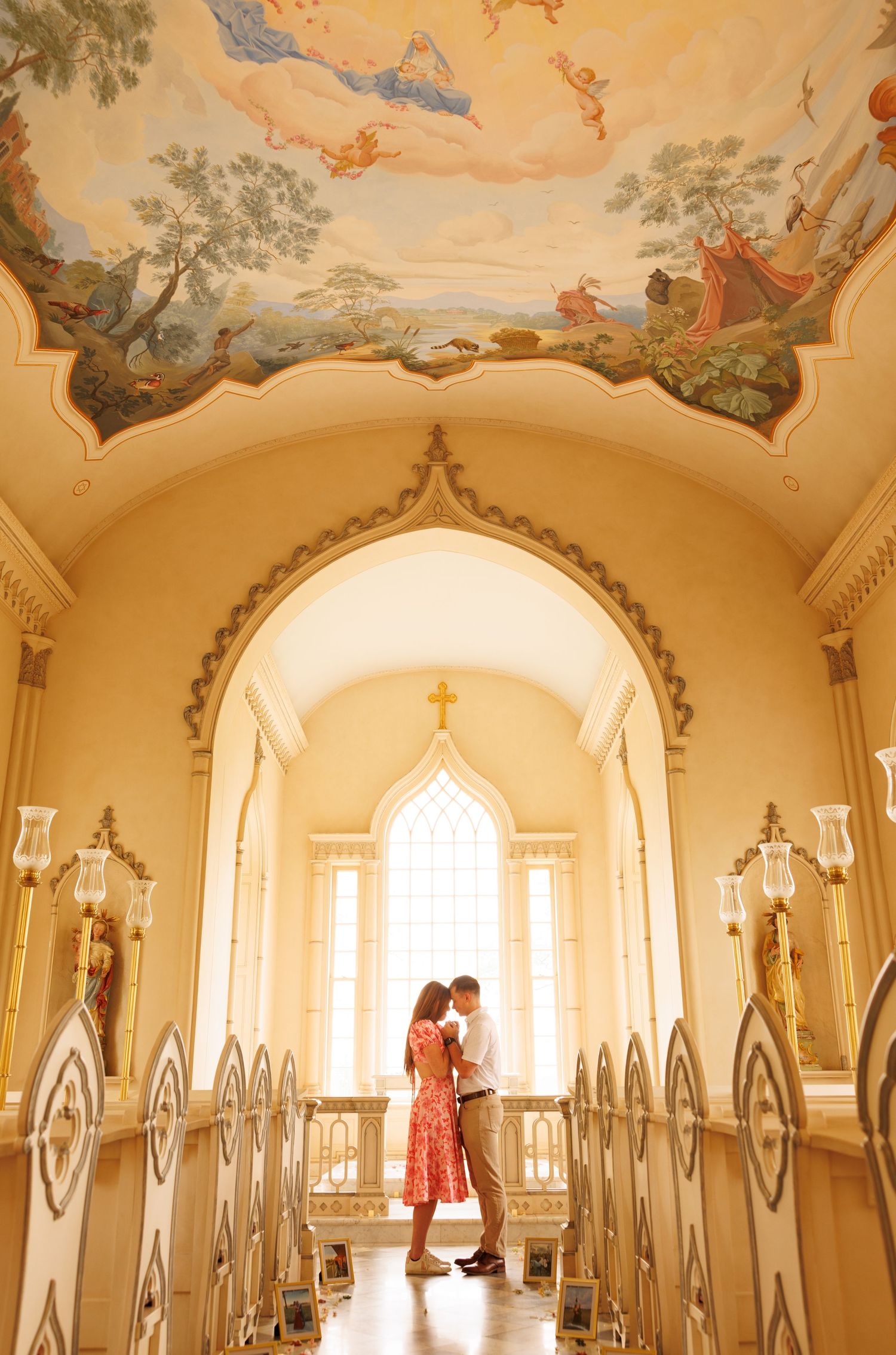 Romantic chapel interior with painted ceiling, arched windows and decorative white railings in soft golden light.