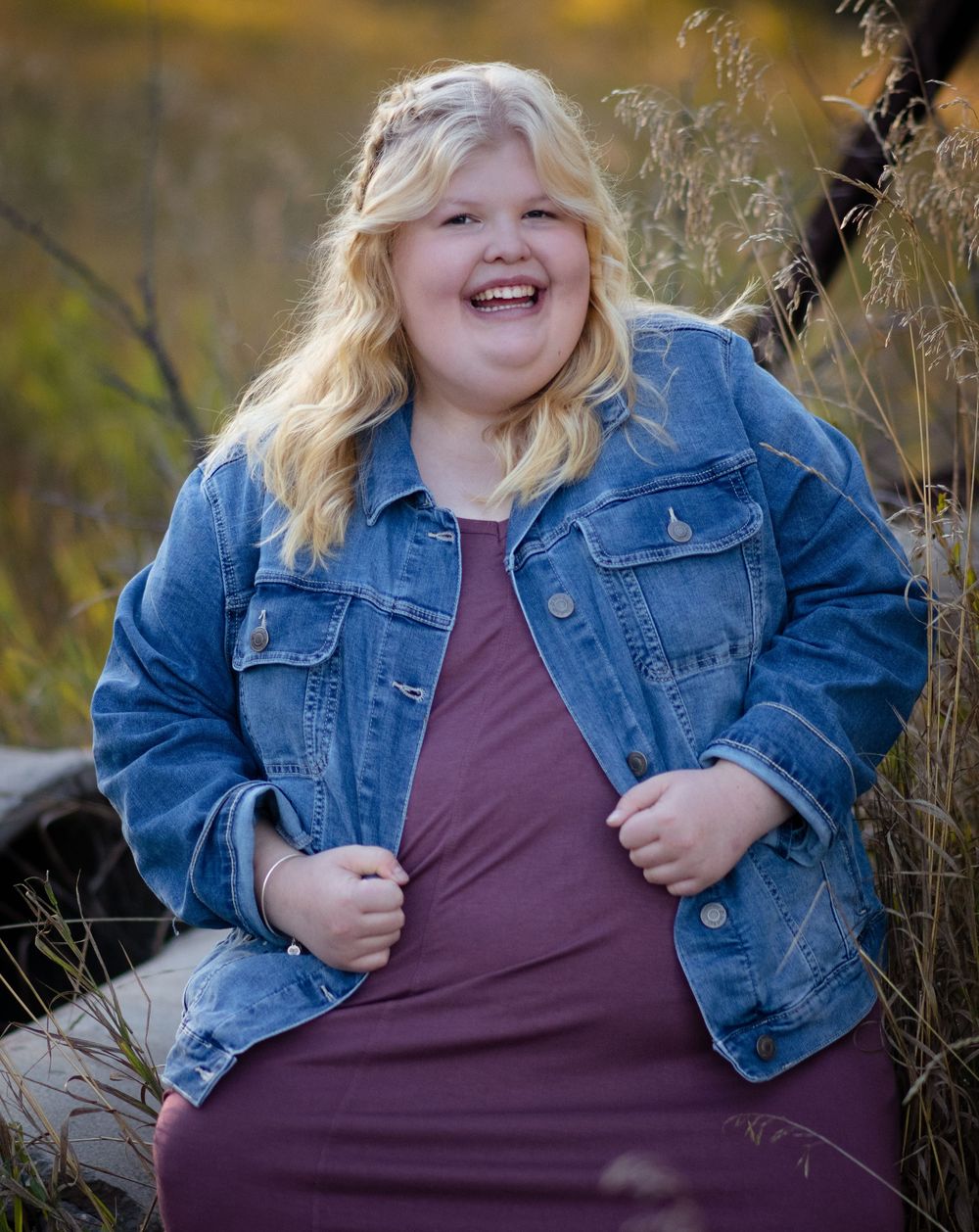 Person wearing denim jacket and purple dress smiles joyfully while posing outdoors among tall grasses in natural lighting.