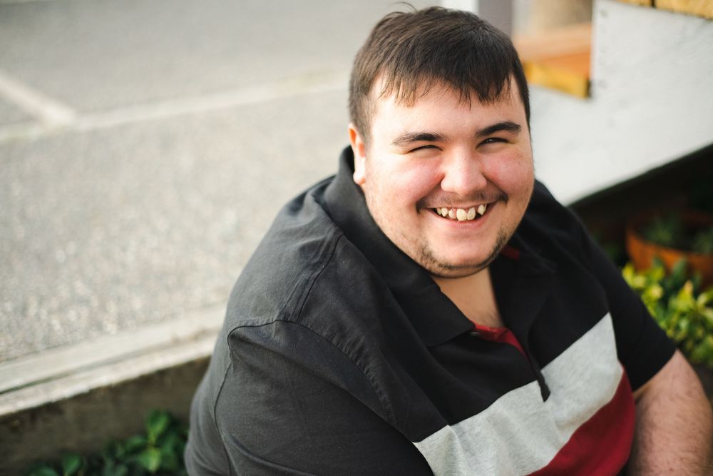 A smiling person wearing a black shirt sits outdoors on a sunny day.