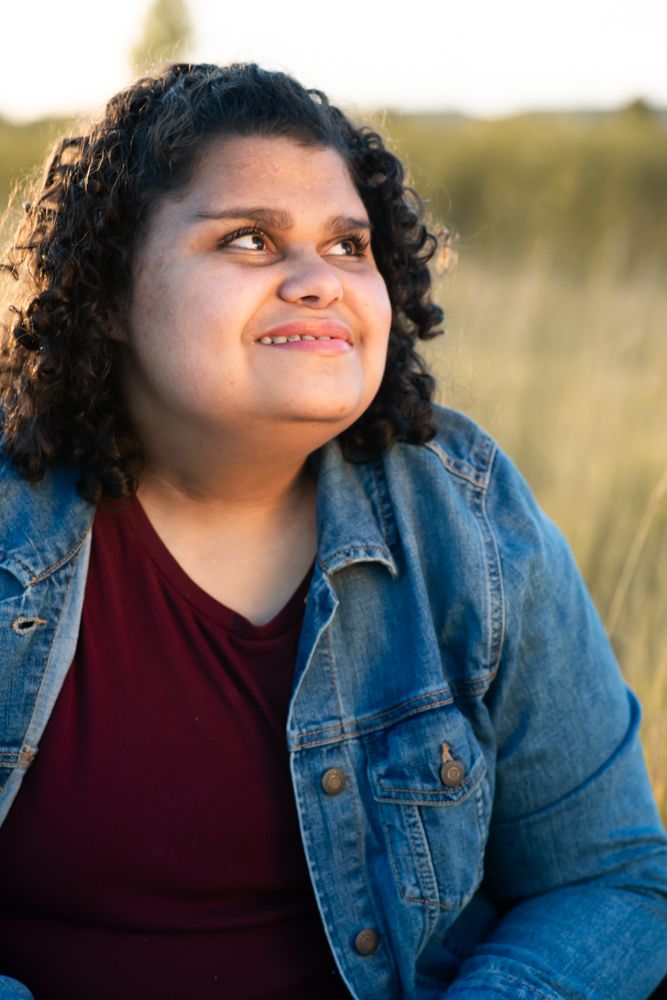 Light shines on someone wearing a denim jacket and burgundy top sitting outdoors in a natural field setting.