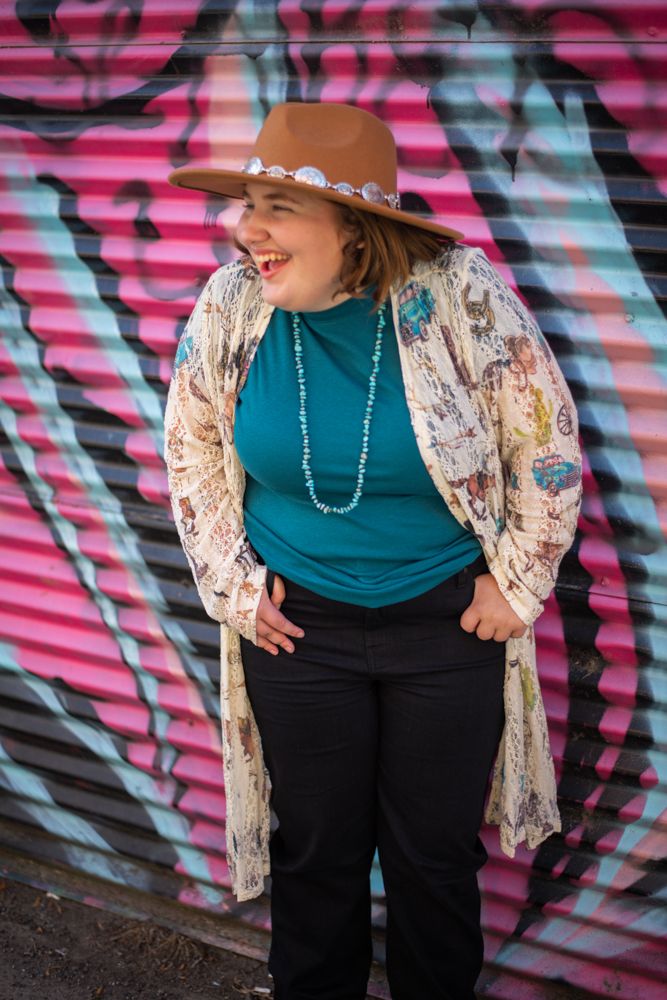 Person in a brown hat and floral kimono poses joyfully against colorful graffiti wall wearing teal top and black pants.