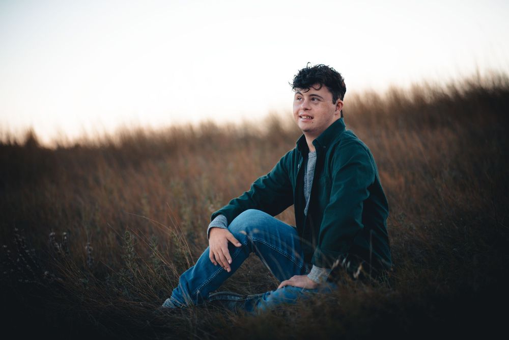 Someone wearing a dark jacket and jeans sits in tall grass during golden hour with a moody sunset backdrop.