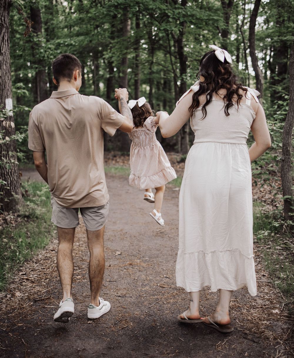 A family walks together holding hands on a forest path while their small child dances in the middle wearing a pink dress.