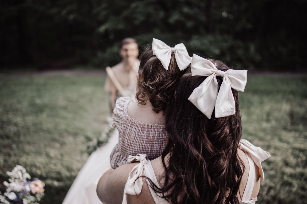 White bows adorn dark brown curled hair at an outdoor wedding celebration with blurred background and flowers.