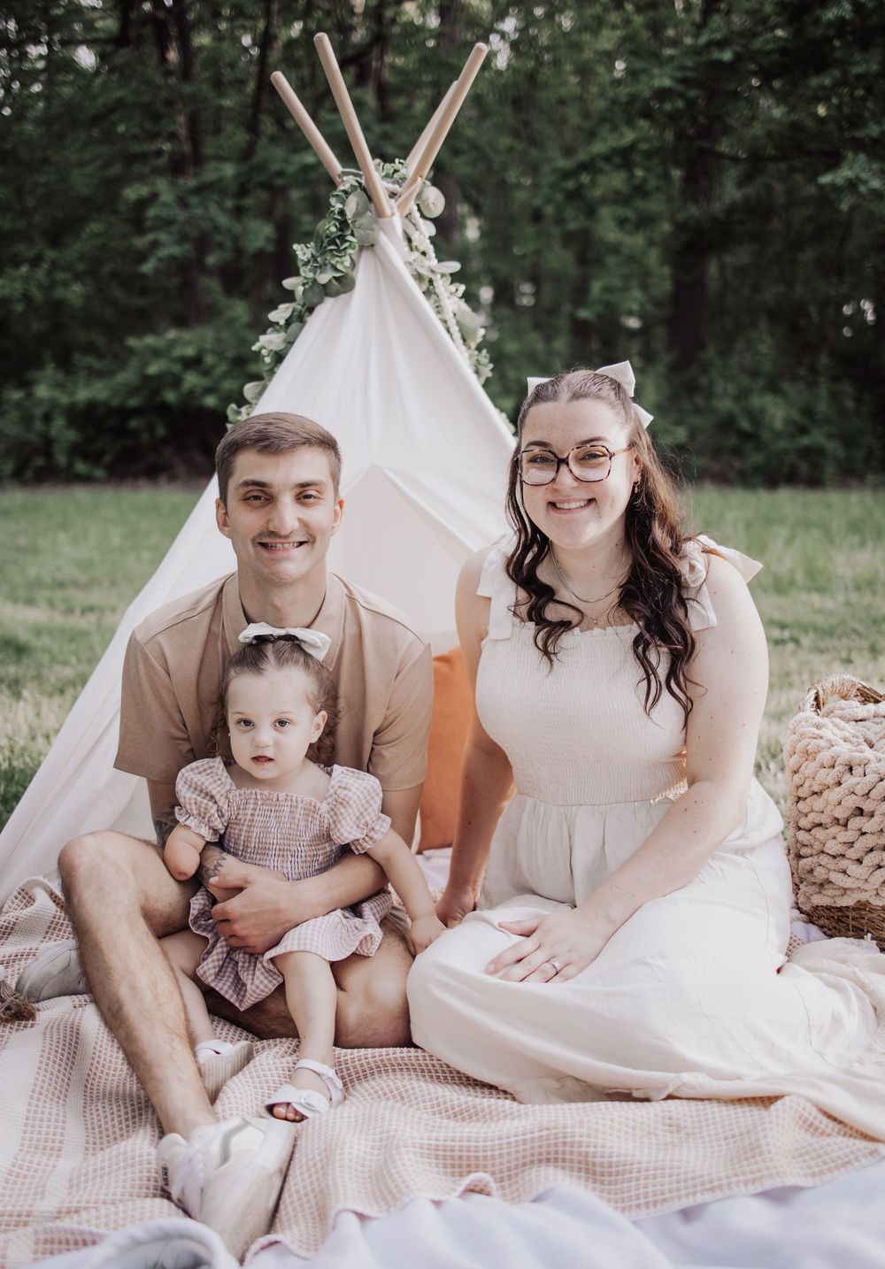 Family enjoys outdoor photoshoot by white teepee tent with florals and blankets on a summer day.