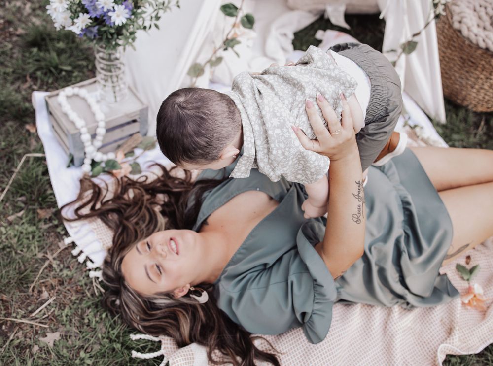 A person in a blue dress lies on a blanket outdoors while playfully lifting a baby wearing a light colored outfit overhead.