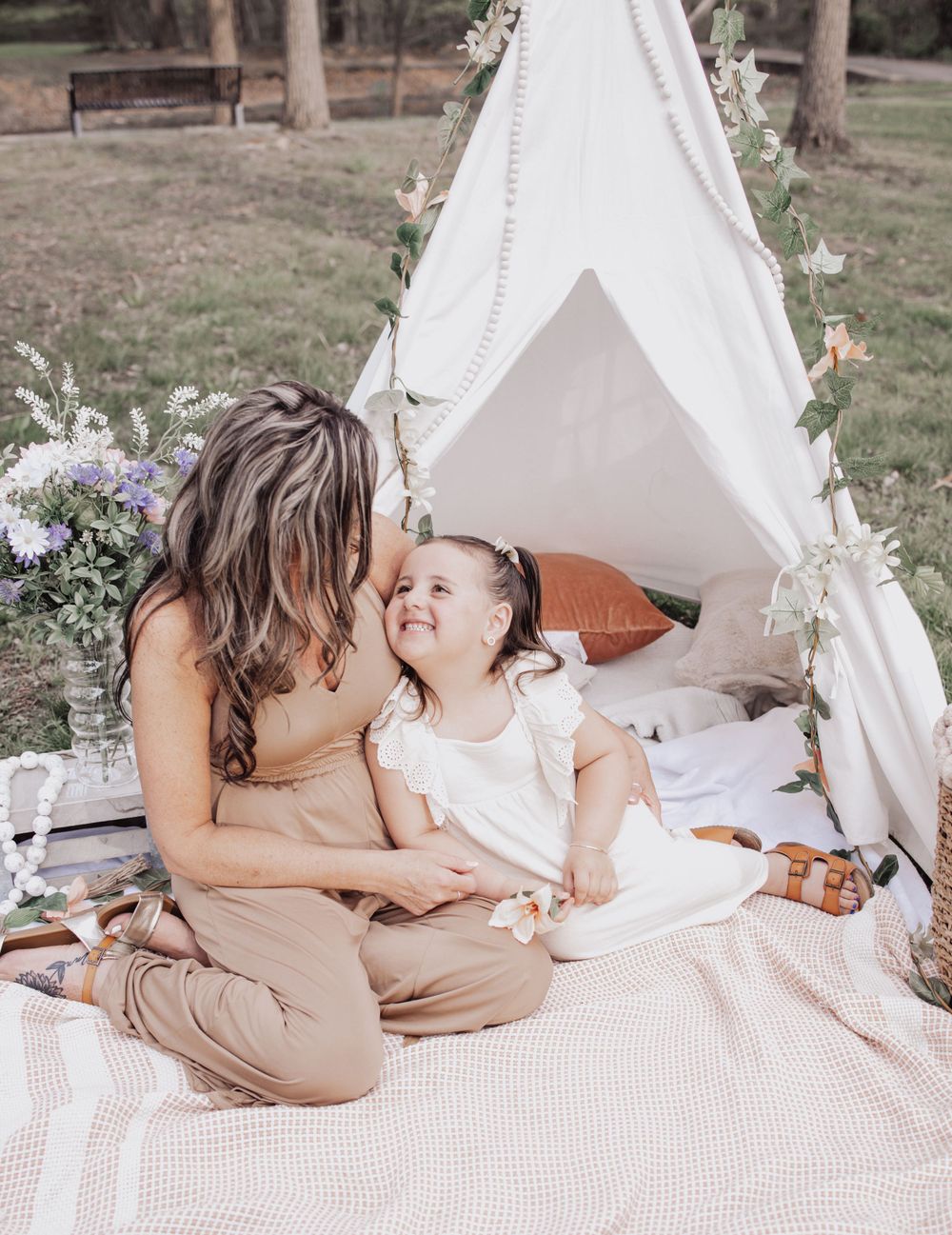 Cozy boho picnic setup with teepee tent, blanket and flowers as parent and child share sweet outdoor moment.