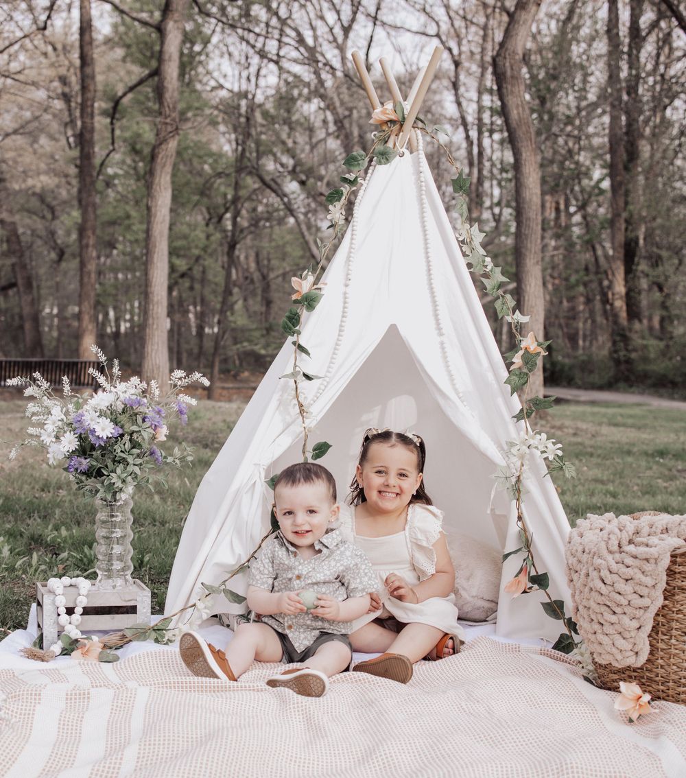 Children sitting in a white teepee tent with floral decorations and blankets during an outdoor photoshoot in a wooded setting.