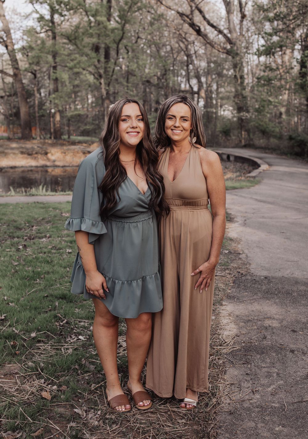 Two women in casual dresses pose together on a wooded path during golden hour in a natural outdoor setting.